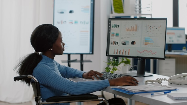 Африканская инвалидная женщина-менеджер-инвалид, работающая за компьютером, показывает данные, обрабатываемые на рабочем столе для проекта, касающиеся экономики, глядя на финансовые документы, проверяя графики, читая рапорты в деловом офисе