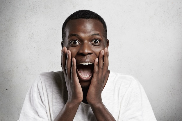 African employee or customer with shocked and surprised face, looking and screaming with big eyes and mouth wide open, holding hands on his cheeks.