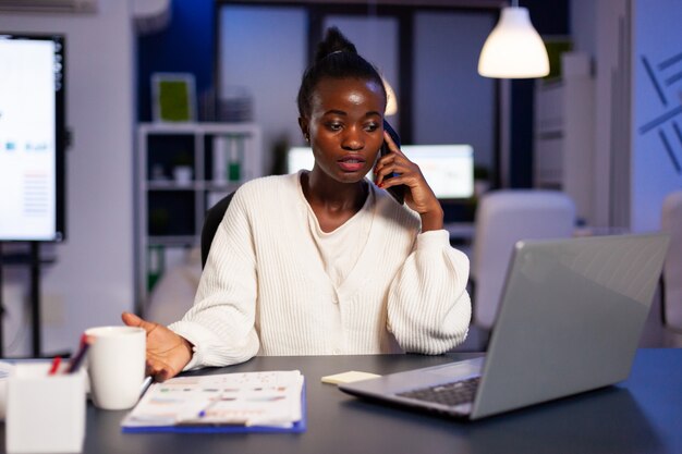 African employee speaking at phone while working at laptop late at night
