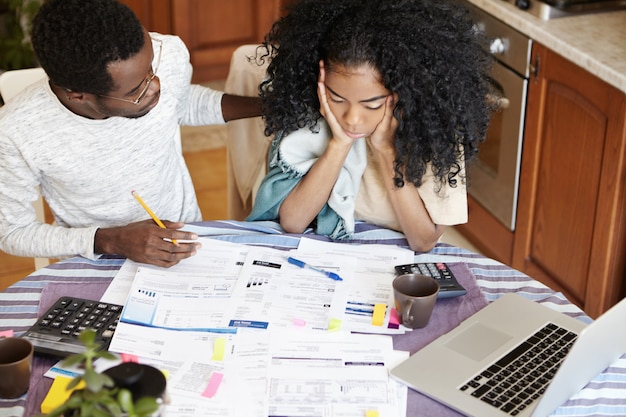 African family doing paperwork together. Stressed young woman holding hands on her face, looking depressed, shocked with amount of family expenses, her supportive husband trying to soothe her