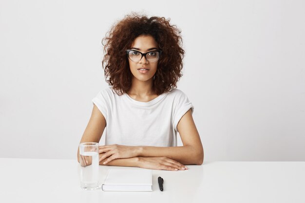 African girl in glasses  sitting over white wall. Copy space.