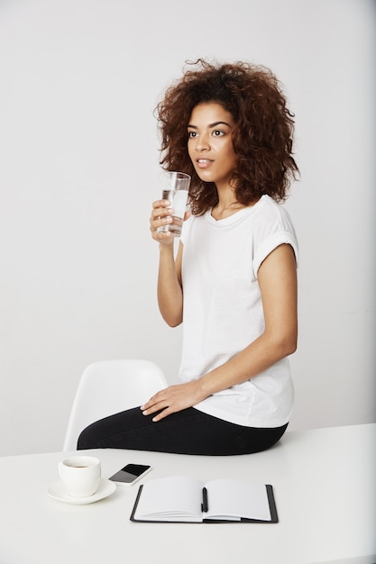 African girl holding glass of water sitting on table over white wall.