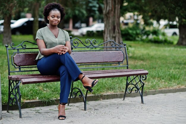 ベンチに座っている緑のブラウスと青いズボンの街のウェアのストリートでポーズをとったアフリカの女の子