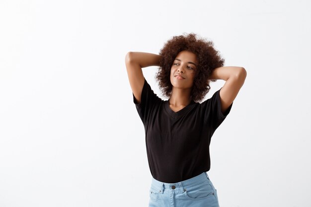 African girl smiling with hands behind head over light wall.