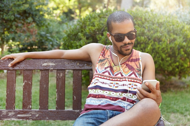 Африканский парень в наушниках сидит на скамейке в городском парке, слушает музыку на своем смартфоне, проверяет электронную почту с помощью мобильного телефона с Интернетом, любит записи и оставляет комментарии в социальных сетях