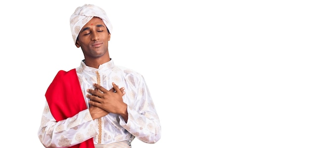African handsome man wearing tradition sherwani saree clothes smiling with hands on chest with closed eyes and grateful gesture on face health concept