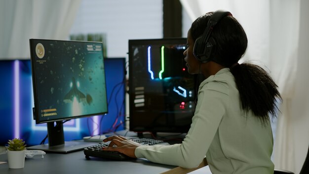 African happy gamer winning space shooter game in gaming home room raising hands. Cyber performing on RGB powerful computer streaming videogames using professional headphones for online championship