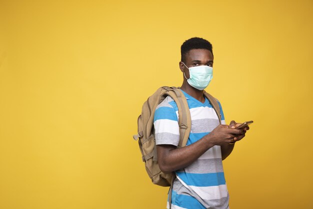 African male with a backpack wearing a facemask and using his phone against a yellow background