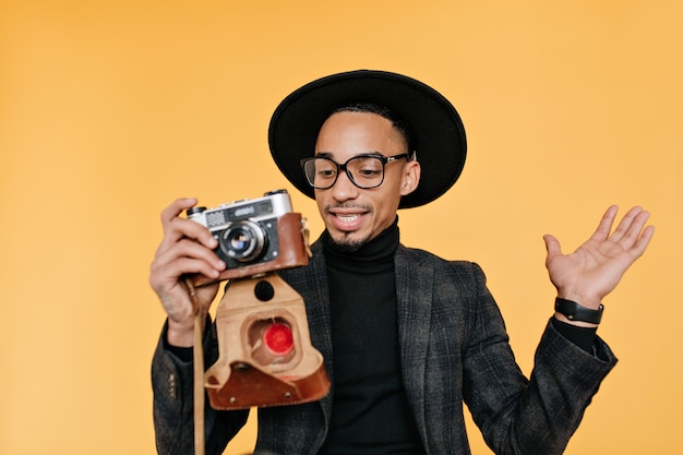 Free photo african man in hat and suit holding camera and expressing amazement.  portrait of carefree black guy posing on yellow wall during photoshoot.