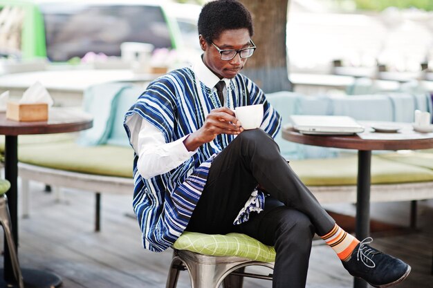 Африканский мужчина в традиционной одежде и очках сидит в уличном кафе, пьет кофе и смотрит на мобильный телефон