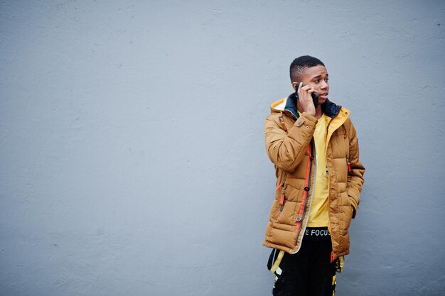 Африканский мужчина в куртке в холодную погоду позирует на улице у серой стены и разговаривает по мобильному телефону
