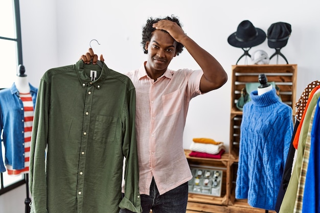 African man with curly hair holding shirt from clothing rack at retail shop stressed and frustrated with hand on head surprised and angry face