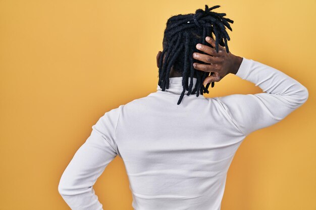 African man with dreadlocks wearing turtleneck sweater over yellow background backwards thinking about doubt with hand on head