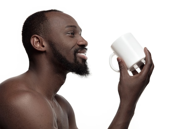 L'uomo africano con bianco tazza di tè o caffè, isolato su bianco di sfondo per studio. chiuda sul ritratto in stile minimalista di un giovane uomo afro felice nudo