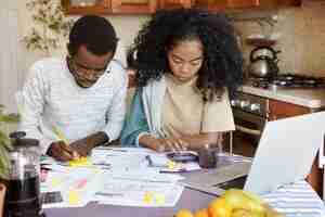 Free photo african man and woman sitting at kitchen table with papers and laptop pc, managing domestic finances together: wife counting on calculator while husband making notes with pencil. family budget