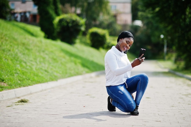 검은 히잡 흰색 스웨트셔츠와 청바지를 입은 아프리카 이슬람 소녀는 휴대전화를 들고 야외에 앉았다