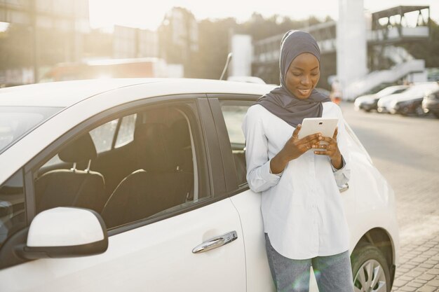 African muslim woman leaning on her car and holding a digital tablet. Working remotely or sharing info.