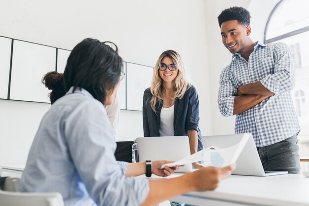 African office worker in checkered shirt standing with arms crossed and looking at asian manager. Indoor portrait of freelance web-developers discussing something and using laptops.