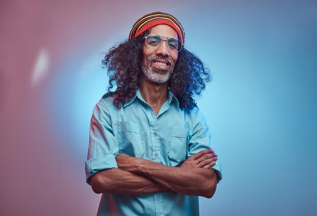 African Rastafarian male smiles and looks at the camera standing with his arms crossed. Studio portrait on a blue background.