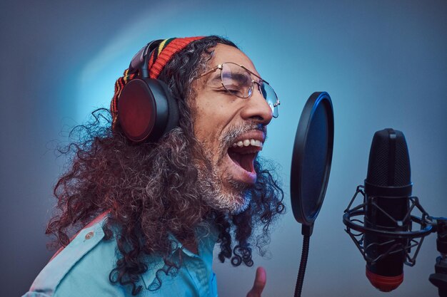 African Rastafarian singer male wearing a blue shirt and beanie emotionally writing song in the recording studio. Isolated on a blue background.