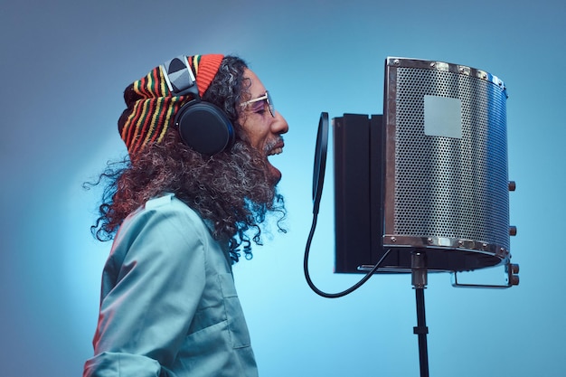 Африканский растафарианский певец в синей рубашке и шапочке эмоционально пишет песню в студии звукозаписи. Изолированные на синем фоне.
