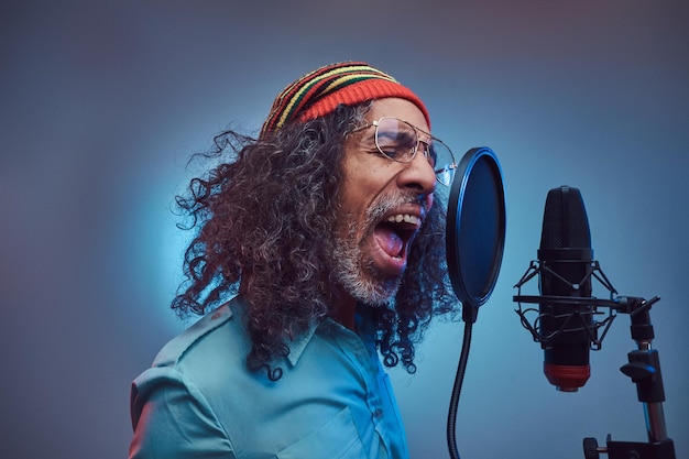 녹음 스튜디오에서 파란색 셔츠와 비니를 입고 감정적으로 노래를 쓰는 아프리카 라스타파리안 가수 남성. 파란색 배경에 고립.