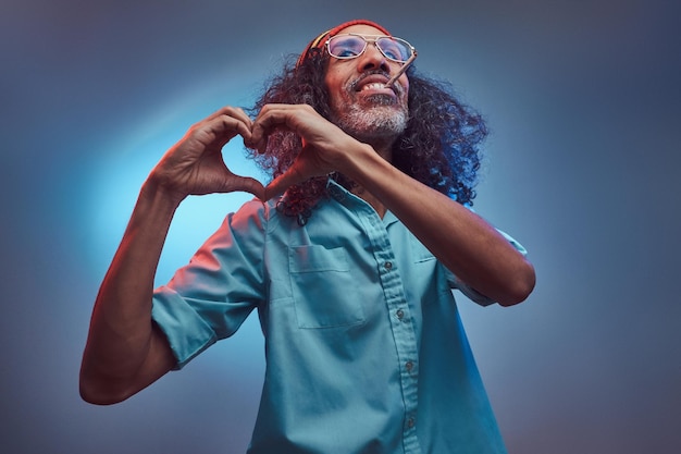 Free photo african rastafarian smokes weed and showing heart symbol and shape with hands. isolated on a blue background.