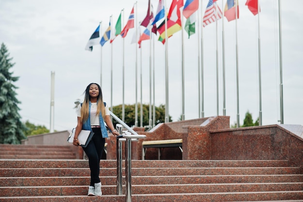 さまざまな国の旗に対して大学の庭でバックパックと学校のアイテムでポーズをとったアフリカの学生の女性