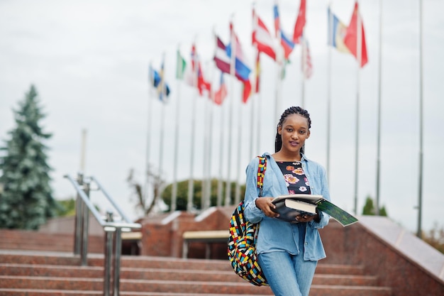 さまざまな国の旗に対して大学の庭でバックパックと学校のアイテムでポーズをとったアフリカの学生の女性