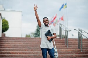 african student male posed with backpack and school items on yard of university against flags of different countries show two fingers