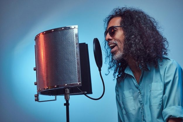 Free photo african vocal artist sings a song loudly next to a microphone condenser. isolated on a blue background.