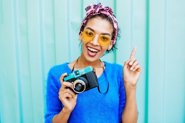 African  woman in bright wool sweater and colorful  headband on hairs holding  film camera.