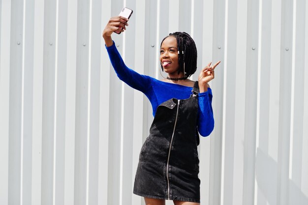 African woman in denim overalls skirt and blue dress posed against white steel wall and making selfie on phone