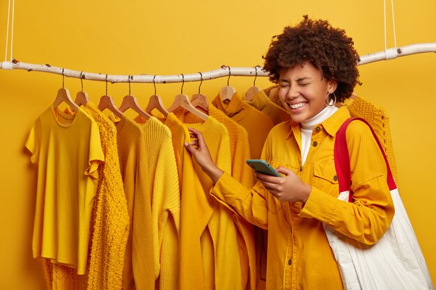 African woman dressed in stylish yellow jacket, carries shopping bag, uses mobile phone for online communication, poses near clothing rail against yellow background