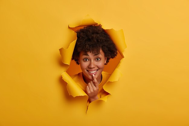 African woman has broad smile, expresses optimism, shows white teeth, holds hand on chin, shares positive memories, poses in ripped hole of yellow background