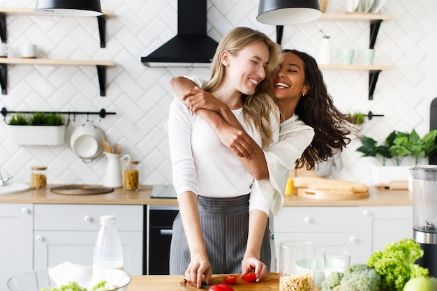 African woman hugging european woman, they stand in the kitchen and laughing