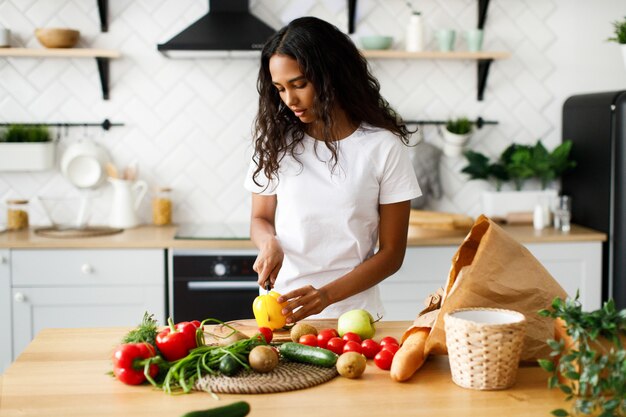 African woman is cutting a yellow pepper on the kitchen desk and on the table are products from a supermarket