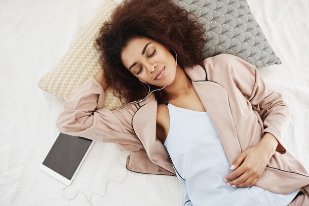 african woman in sleepwear smiling listening to music in headphones lying on bed.