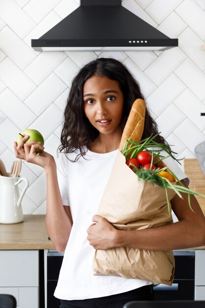 African woman stands on the kitchen holding a paper bag with groceries and has surprised look