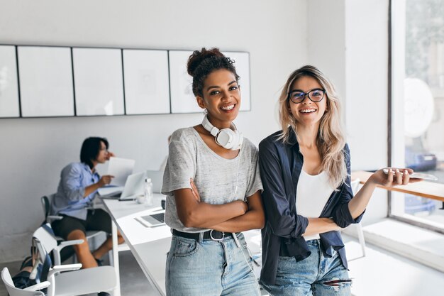 African woman in trendy casual attire came to friend's office and posing with asian programmer. Charming blonde secretary having fun during break while her japanese colleague working.