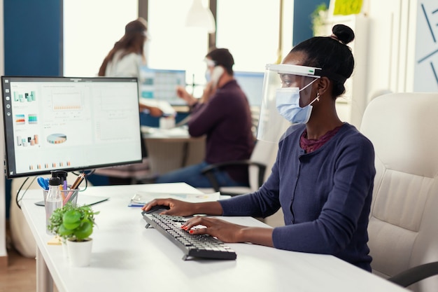 African woman typing on computer at workplace wearing face mask as safety precaution agasinst covid19