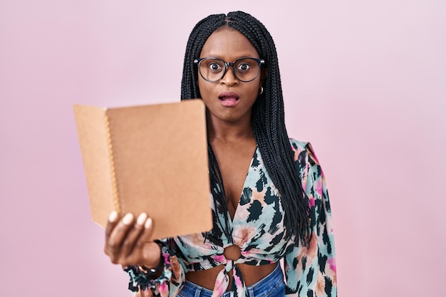 African woman with braids reading a book scared and amazed with open mouth for surprise, disbelief face