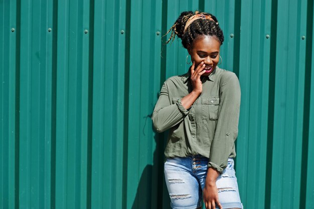 African woman with dreads hair in jeans shorts posed against green steel wall