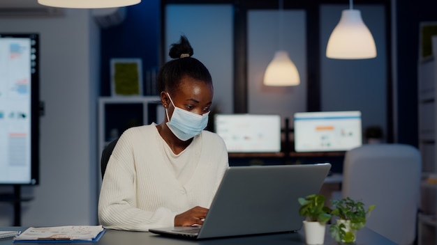 African woman with face mask reading emails late at night to respect deadline of project working in new normal business office, analysing documents, making strategy overtime during global pandemic