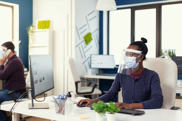 安全対策としてcovid19に対してフェイスマスクを着用して職場で働くアフリカ人。新しい通常のビジネス金融オフィスの多民族チームがレポートをチェックし、デスクトップを見てデータを分析します。新しい