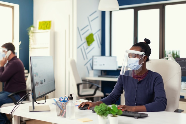 African working at workplace wearing face mask against covid19 as safety precaution. Multiethnic team in new normal business financial office checking reports, analysing datas looking at desktop. New