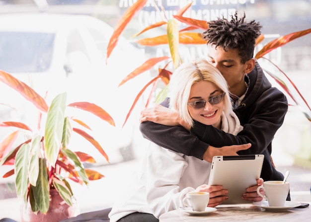 An african young man loving her smiling girlfriend sitting in cafe looking at digital tablet