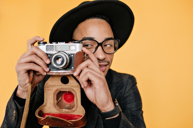 African young man playfully posing with camera on yellow wall. Indoor photo of talented photographer in elegant hat having fun