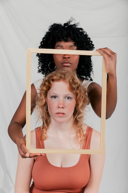 Free photo an african young woman holding wooden frame in front of caucasian female against grey backdrop