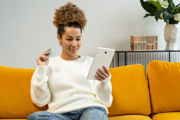 홈쇼핑에서 태블릿 컴퓨터와 신용카드를 들고 소파에 앉아 있는 아프리카계 미국인 여성